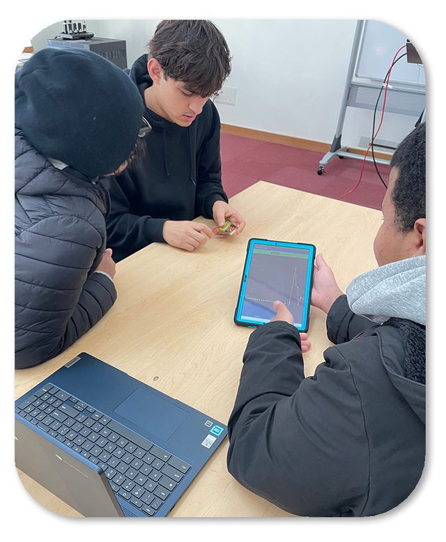Three students around a table: one holds a Micro:bit while another watches its readings graphed on a tablet.