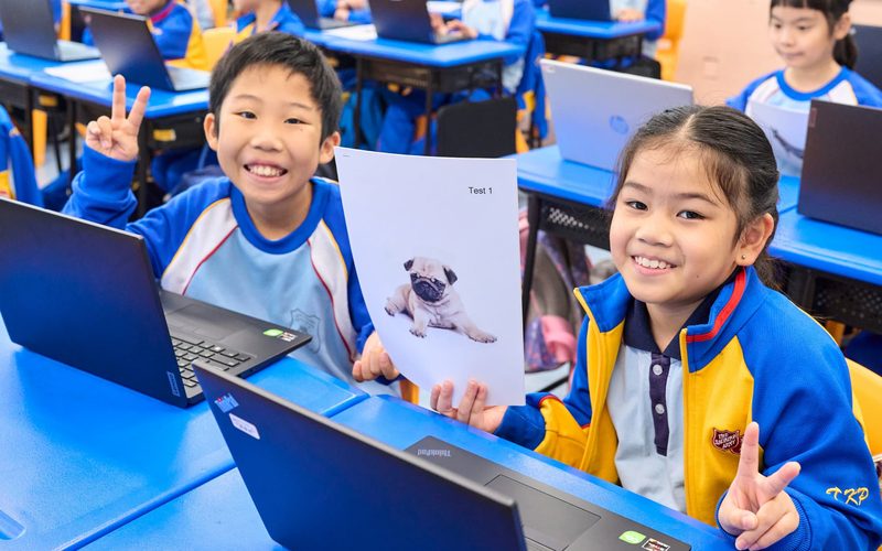 Two smiling primary school students at their laptops, one holding up a printed picture of a pug, in a classroom full of students working on laptops.