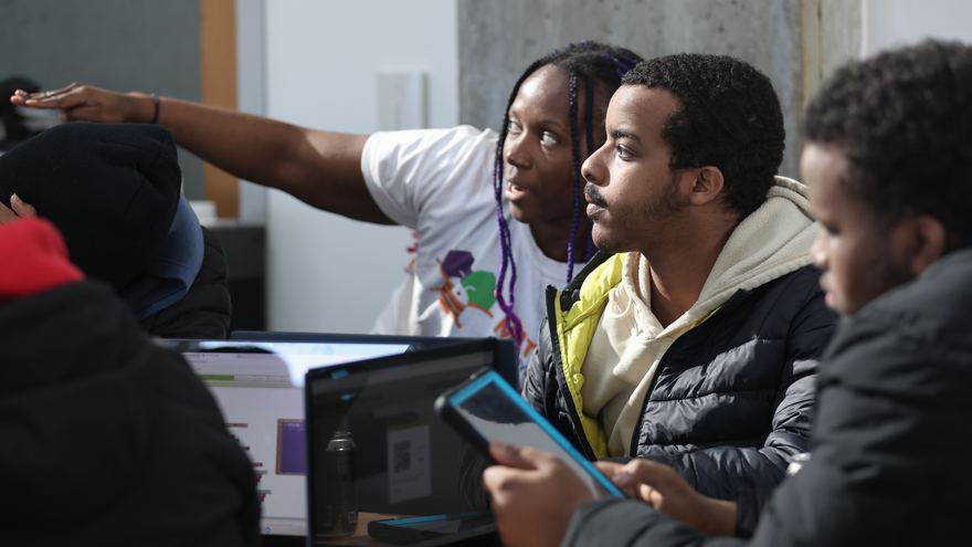 An MIT App Inventor instructor points something out to high school students working on laptops and a tablet.