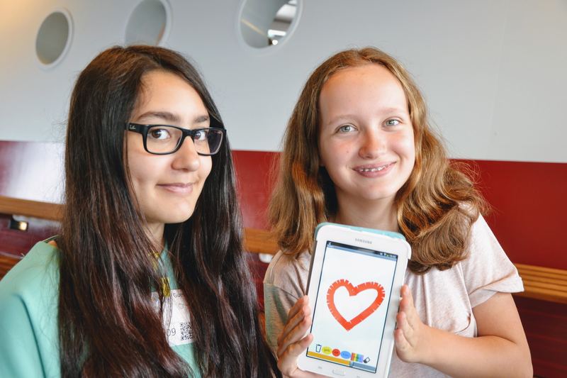 Two students smiling at the camera, one holding up a phone running the heart-drawing app they built.