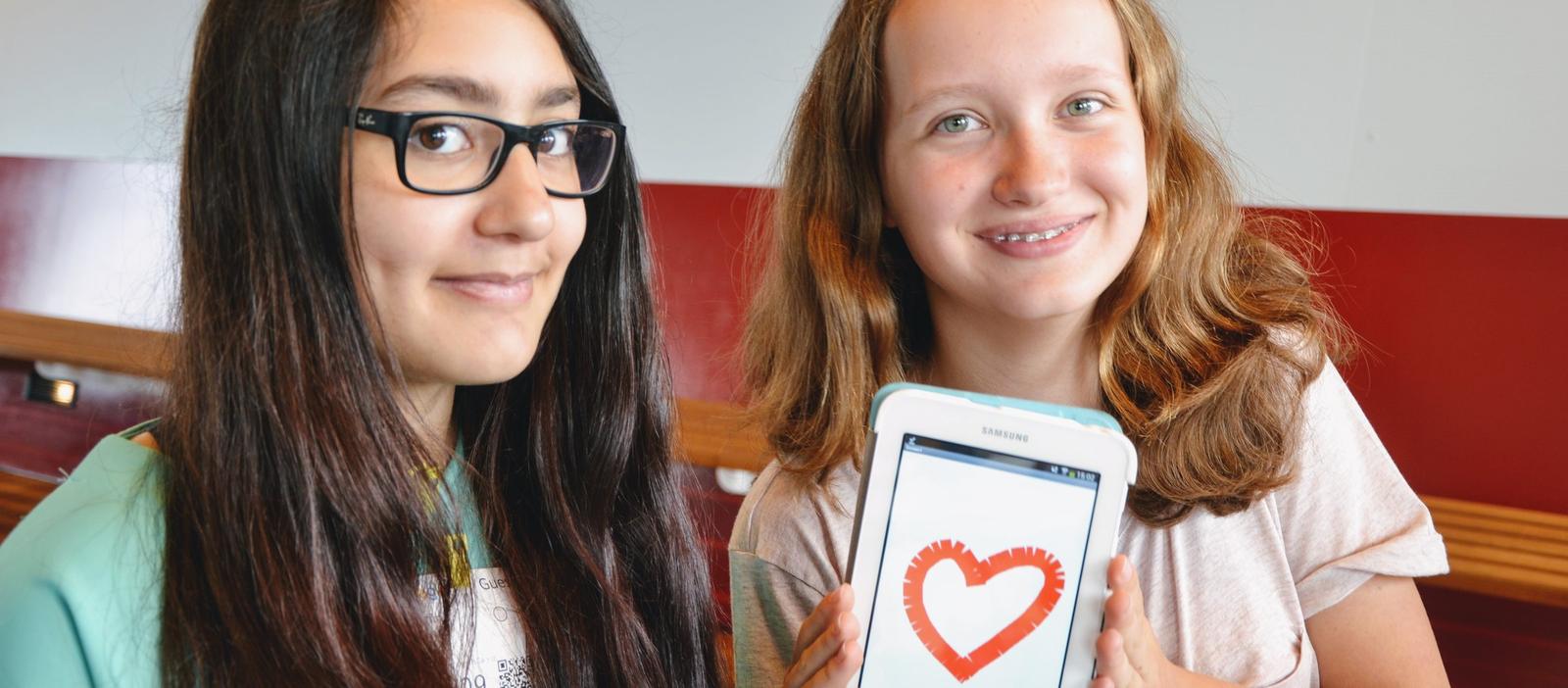 Two students smiling at the camera, one holding up a phone running the heart-drawing app they built.